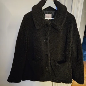 Svart teddyjacka  - Nyskick Säljer en supermysig svart teddyjacka. Den har en fluffig textur och stängs med knappar framtill. Jackan har långa ärmar och en innerficka med dragkedja. Perfekt för kyliga höst- och vinterdagar!