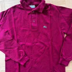 Vinröd pikétröja från Lacoste - Säljer en snygg vinröd pikétröja från Lacoste. Den har långa ärmar och klassisk krage med knappar. Perfekt för både vardag och lite finare tillfällen. Tröjan är i bra skick och redo för en ny ägare!
