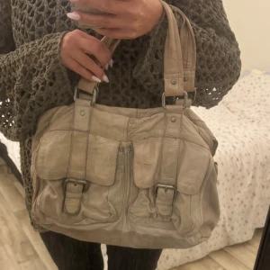 Beige axelväska i skinn - Säljer en snygg beige axelväska i skinn med praktiska fickor och spännen framtill. Väskan har en dragkedja upptill och är rymlig med flera fack inuti. Ser lite platt ut på bilden då den är tom men med saker i ser den mycket bra ut!