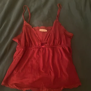 Röd linne från Nelly - Säljer en superfin röd linne från Nelly. Den har smala axelband och en söt liten rosett framtill. Toppen är dekorerad med spetsdetaljer längs kanterna och har en v-ringad design. Perfekt för sommardagar eller en mysig hemmakväll💕