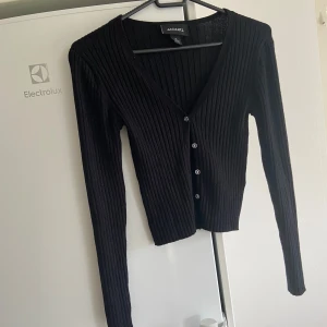 Svart ribbad kofta från Monki - Säljer en snygg svart ribbad liten kofta från Monki i storlek XS i väldigt bra skick. Den har en v-ringad design med glittriga blommiga knappar framtill. Perfekt för både vardag och fest! 🖤alla kläder är tvättade men som legat i förrådet. 