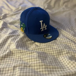 Blå keps med LA-broderi - Säljer en snygg blå keps från New Era med ett klassiskt LA-broderi i vitt framtill. Kepsen har också ett coolt broderat motiv av palmer och text på sidan. Perfekt för att ge din outfit en sportig touch!