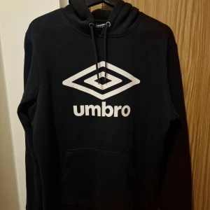 Svart hoodie från Umbro - Säljer en svart hoodie från Umbro i storlek L. Den är superbekväm och perfekt för både vardag och träning. Hoodien har en klassisk design med Umbro-loggan i vitt på framsidan och en praktisk magficka. Materialet är en blandning av bomull och polyester, vilket gör den både mjuk och hållbar. Perfekt för höst och vinter!