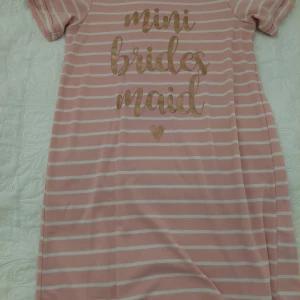 Rosa randig klänning med text - Söt rosa nattlinne med vita ränder och texten 'mini bridesmaid' i guld. Perfekt för en liten tärna på bröllopet! Klänningen har korta ärmar och är gjord i ett mjukt och bekvämt material. Det skönaste nattlinnet.