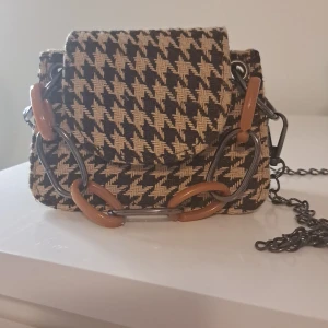 Rutig axelväska med kedjedetaljer - Säljer en supercool axelväska med ett klassiskt rutigt mönster i svart och beige. Väskan har en unik kedja med stora länkar i brunt och metall, vilket ger den en edgy look. 100% nytt och oanvänd. Den är mini väskan. Köpte den för 150. Utan kjedian.