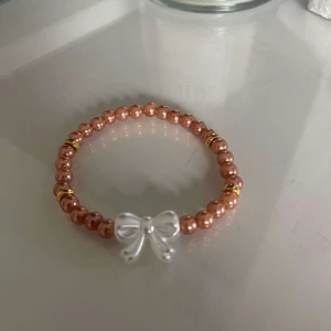 Armband med pärlor och rosett - Säljer ett supergulligt armband med skimrande pärlor i en varm kopparfärg. Armbandet har små guldfärgade detaljer mellan pärlorna och en söt silverfärgad rosett som pricken över i:et. Perfekt för att piffa upp vilken outfit som helst!