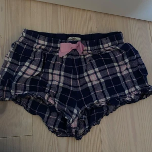 Rutig shorts med rosa rosett - Superfina rutiga pyjamasshorts i mörkblått och rosa med en söt rosa rosett framtill. Perfekta för en mysig hemmakväll. Köpta på Kappahl och är i stl 158/164 men passar även xs☺️