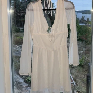 Vit klänning från Divided - Säljer en superfin vit klänning från Divided. Den har långa ärmar och en snygg v-ringad design med knytning framtill. Perfekt för sommarens fester eller en dag på stan. Klänningen är i nyskick och gjord av ett lätt och luftigt material.