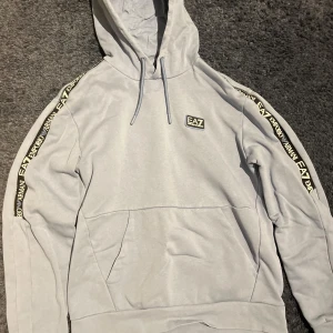 Blå hoodie från Emporio Armani - Snygg grå hoodie från Emporio Armani med EA7-logga på bröstet och längs ärmarna. Den har en klassisk känguruficka och justerbar huva med dragsko. Perfekt för en stilren och bekväm look, passar nån runt 150-162,prick kan diskuteras 