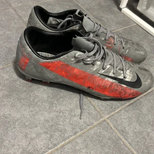 Grå och röda fotbollsskor från Nike - Snygga grå fotbollsskor från Nike med röda och svarta detaljer. Skorna har snörning och är designade för optimal prestanda på planen. Perfekta för fotbollsentusiaster som vill ha stil och funktionalitet
