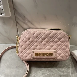 Rosa Moschino väska  - Jätte fin rosa axelväska från Love Moschino, väskan har massa små fina guld detaljer samt flera fack inuti. Väskan är helt ny och aldrig använd. Original påsen medföljer, kontakta mig vid fler bilder eller frågor. Såklart äkta‼️