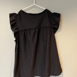 Svart blus med spetsdetaljer - Säljer en svart blus med spetsdetaljer och korta ärmar. Blusen har en luftig passform och ett diskret hålmönster som ger en elegant touch. Perfekt för både vardag och speciella tillfällen.💕