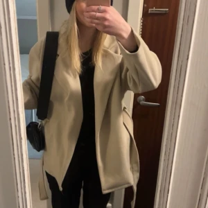 Beige kappa med bälte - Säljer en stilren beige kappa med bälte. Jackan har en elegant design med långa ärmar och en lös passform. Perfekt för höst och vår. En klassisk och tidlös look som funkar till många tillfällen.