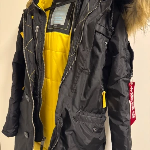 Svart jacka med pälsdetaljer från Alpha Industries - Säljer en cool svart jacka från Alpha Industries med gul insida och pälsdetaljer på huvan. Jackan har dragkedja och knappar, samt en ikonisk 'Remove Before Flight'-tag. Perfekt för kyliga dagar med stil. Har använt två månader 
