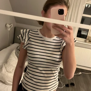 Randig topp från Holly & Whyte - Säljer en snygg randig topp från Holly & Whyte by Lindex. Den är vit med svarta ränder och har korta ärmar med volangdetaljer. Perfekt för en stilren look!