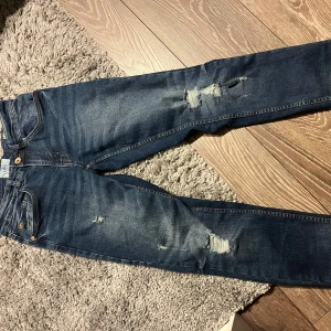 Blå jeans med slitningar  - Snygga blå jeans med slitna detaljer på framsidan. De har en klassisk femficksdesign och knappgylf. Perfekta för en avslappnad stil. Köpte dessa på Plick för 200kr, men passade tyvärr inte mig