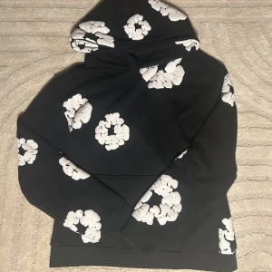 Svart hoodie med vita blommor från Denim Tears - Säljer en svart hoodie från Denim Tears med stora vita blommor som mönster. Den har en klassisk huva och är perfekt för en avslappnad stil. Passar bra till både jeans och joggers.