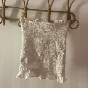 Vit smockad tubtopp - Säljer en söt vit smockad tubtopp med volangdetaljer upptill och nedtill. Perfekt för sommardagar och kan enkelt matchas med olika outfits. Toppen är elastisk och bekväm att bära.