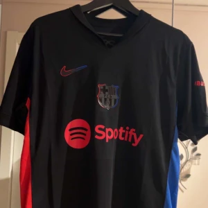 Svart Barcelona tröja  - Snygg svart Barca fotbollströja från Nike med röda och blå detaljer. Tröjan har korta ärmar och klubbmärke på bröstet. Perfekt för fotbollsfans som vill ha en stilren look. Tröjan är helt oanvänd och har till och med taggsen kvar. 