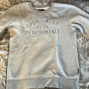 Grå sweatshirt från Peak Performance - Säljer en grå sweatshirt från Peak Performance med texten 'SPORTSWEAR PEAK PERFORMANCE' på framsidan. Tröjan är tillverkad i en mjuk bomullsblandning och har långa ärmar samt ribbade muddar vid ärmslut och nederkant. 