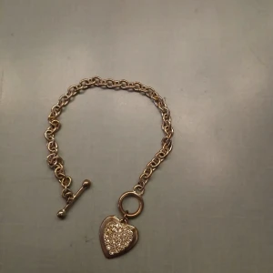 Silver armband med hjärthänge - Elegant silverarmband med ett hjärthänge dekorerat med glittrande stenar. Armbandet har en klassisk kedjedesign och stängs med en stavlås. Perfekt för att ge en touch av glamour till din stil.