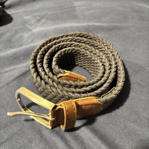  Onewear Flätat bälte - Snyggt flätat bälte i grått med silverfärgat spänne och bruna läderdetaljer. Perfekt för att ge en stilren touch till din outfit. Bältet är flexibelt och passar de flesta storlekar.