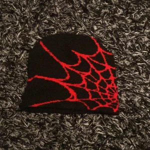 Svart mössa med rött spindelnätsmönster - Cool svart mössa med ett iögonfallande rött spindelnätsmönster. Perfekt för att ge en edgy touch till din outfit. Passar bra för kyligare dagar.