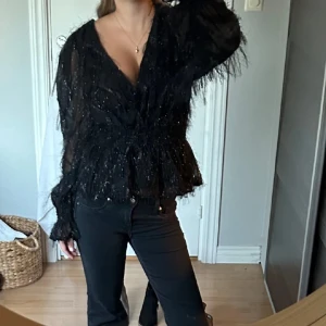 Svart glittrig blus - Säljer en elegant svart blus med glittriga detaljer. Blusen har långa ärmar och en v-ringad design med knytning i midjan. Perfekt för en kväll ute eller en speciell tillställning.✨