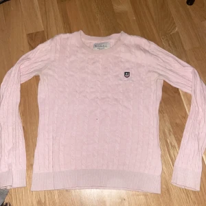 Rosa stickad tröja från Bondelid - Säljer en söt rosa stickad tröja från Bondelid med kabelstickat mönster och broderad logga på bröstet. Tröjan har långa ärmar och rund halsringning. Perfekt för en mysig dag!