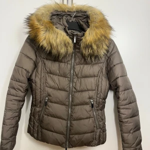 Militärgrön dunjacka med pälsdetalj från Livelo - Säljer en snygg militärgrön dunjacka från Livelo med en fluffig pälsdetalj på huvan. Jackan har dragkedja framtill och två praktiska fickor med dragkedjor. Perfekt för kyliga dagar!