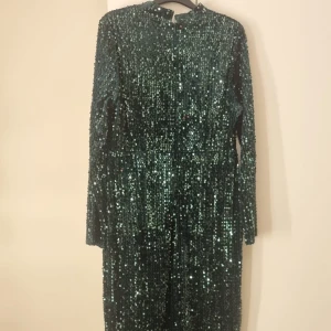 Grön glittrig långklänning - Fantastisk grön långklänning täckt av glittrande paljetter. Klänningen har långa ärmar och en elegant volangdetalj vid fållen. Perfekt för att göra ett statement på festen!