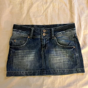 Jeanskjol från Cada Dia - Snygg jeanskjol från Cada Dia i mörkblå denim med dekorativa nitar på bakfickorna. Kjolen har en klassisk femficksdesign och stängs med två knappar framtill. 