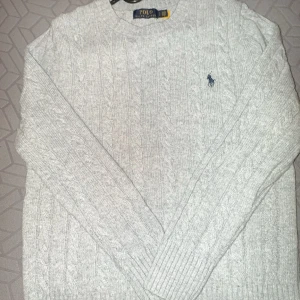 Grå stickad tröja från Ralph Lauren - Säljer en stilren grå stickad tröja från Ralph Lauren med kabelstickat mönster. Tröjan har långa ärmar och ribbstickade muddar vid ärmslut och nederkant. Perfekt för kyligare dagar!