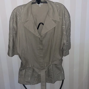 Beigegrå  kavaj & byxor  - Snygg beigegrå sommar kavaj & byxor  med knappar och bälte i midjan i linne. Den har trekvartsärmar och ett diskret broderat mönster som ger en elegant touch.Kavaj st.40 och byxor st.38. Perfekt för att lyfta vilken outfit som helst.