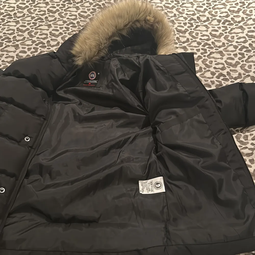 Säljer en svart dunjacka från Canada Goose med pälsdetaljer på huvan. Jackan har en klassisk design med lång ärm och är perfekt för kalla dagar. Den har en dragkedja framtill och är märkt med Canada Goose-logotypen på ärmen.. Takit.