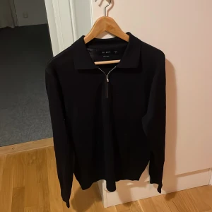 Svart tröja med dragkedja från Riley - Snygg svart tröja från Riley med dragkedja framtill. Perfekt för en stilren look. Tröjan har långa ärmar och en klassisk krage.