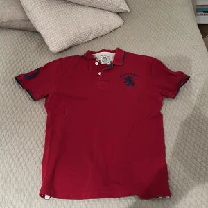 Röd pikétröja från Hackett - Snygg röd pikétröja från Ralph Lauren med broderad logga på bröstet. Tröjan har korta ärmar och en klassisk krage med knappar. Perfekt för en avslappnad stil.