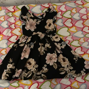 Svart blommig klänning - Charmig svart klänning med stora blommor i ljusa toner. Klänningen har en elegant passform med korta ärmar och en rund halsringning. Perfekt för en somrig look med sitt vackra blommönster.