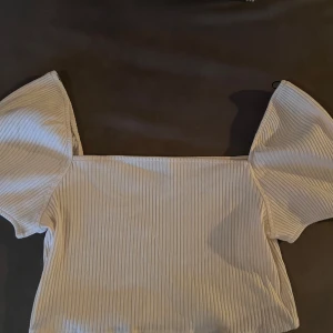 Vit ribbad topp från H&M - Säljer en stilren vit ribbad topp från H&M. Toppen har korta ärmar och en luftig passform, perfekt för varmare dagar. Den är enkel att matcha med olika outfits och ger en fräsch look.