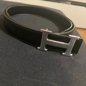 Bälte - Snyggt svart bälte med ett iögonfallande silverfärgat H-spänne. Perfekt för att ge en stilren touch till din outfit. Bältet är tillverkat i skinn och har en klassisk design som passar till många olika stilar.