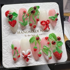 Handgjorda naglar med jordgubbs- och nyckelpigemotiv - Unika handgjorda naglar dekorerade med 3D-motiv av jordgubbar och nyckelpigor. Designen har en mix av rosa och vita basfärger med gröna blad och röda detaljer. Perfekt för en lekfull och somrig look.