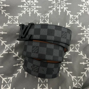 LV bälte svart - Snyggt bälte från Louis Vuitton med klassiskt rutigt mönster i grått och svart. Bältet har en stilren svart spänne med LV-logotypen. Tillverkat i skinn och perfekt för att ge en lyxig touch till din outfit.