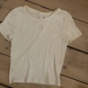 Vit ribbad topp från H&M - Säljer en vit ribbad topp från H&M med korta ärmar och ett hjärtformat hål på framsidan. Perfekt för en avslappnad stil. Toppen är i en mjuk och bekväm bomullsmix.