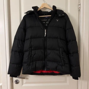 Svart pufferjacka från Gant - Säljer en svart pufferjacka från Gant med dragkedja och broderad logga på bröstet. Jackan har en huva och långa ärmar, perfekt för kyliga dagar. Den är stilren och enkel att matcha med olika outfits.