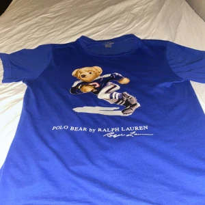 Blå t-shirt med Polo Bear från Ralph Lauren - Säljer en blå t-shirt från Ralph Lauren med en cool Polo Bear-tryck på framsidan. T-shirten har korta ärmar och är perfekt för en avslappnad stil. Den är gjord i mjukt material för extra komfort. Perfekt för dig som gillar sportig design!