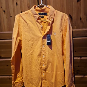Orange skjorta från Polo Ralph Lauren - Snygg orange skjorta från Polo Ralph Lauren i slim fit. Skjortan har långa ärmar och knappar framtill. Den är tillverkad i ett lätt twillmaterial och har en broderad logotyp på bröstet. Perfekt för en stilren look! Aldrig använd innan. Lappen sitter kvar med extra knappar i en liten påse