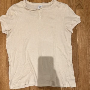 Beige t-shirt från Zara - Säljer en stilren beige t-shirt från Zara med korta ärmar och knappar vid halsen. Perfekt för en avslappnad look.