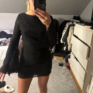 Svart glittrig klänning - Snygg svart glittrig klänning med lång ärm och öppen rygg. Klänningen har en tight passform och knytning i ryggen. Perfekt för en kväll ute eller en festlig tillställning. Använd en gång på nyår! 