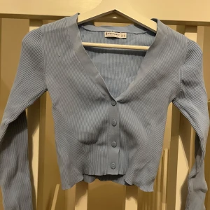 Ljusblå ribbad kofta från Stradivarius - Säljer en ljusblå ribbad kofta från Stradivarius. Koftan har en v-ringad design med knappar framtill och långa ärmar. Perfekt för en stilren och bekväm look.