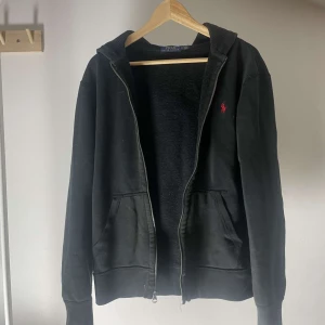 Svart hoodie från Ralph Lauren - Säljer en svart hoodie från Ralph Lauren med dragkedja och huva. Den har en röd broderad logga på bröstet och är tillverkad i ett mjukt material. Perfekt för en avslappnad stil.